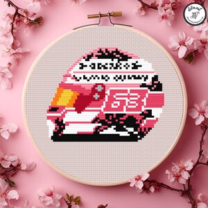 May include: A cross-stitch embroidery of a pink and white racing helmet with the number 58. The design includes black and yellow accents, and text. The embroidery is set in a wooden hoop, surrounded by pink flowers.
