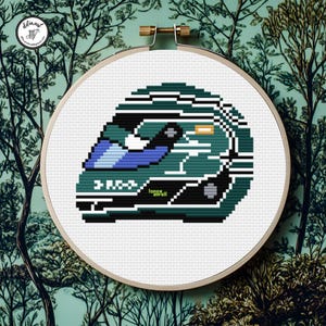 May include: Cross-stitch embroidery of a teal and blue racing helmet, displayed in a wooden hoop. The helmet design includes black and white accents, with the name "Lance Stroll" visible. The background features a teal and white tree pattern.