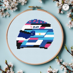May include: A cross-stitch design of a racing helmet, featuring a color scheme of blue, white, pink, and black. The helmet has the text "D.M.T." on the side. The design is set within a wooden embroidery hoop, surrounded by white flowers.