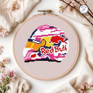 May include: A cross-stitch embroidery piece in a wooden hoop featuring a stylized racing helmet design. The helmet is white with pink, yellow, and dark blue accents, displaying the red and white Red Bull logo and the text Red Bull in red.