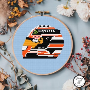 May include: Cross-stitch embroidery of a racing helmet in a wooden hoop. The helmet design features orange, black, and white stripes, with the name "OSCAR" and the word "HALTER" visible. The background is light blue.