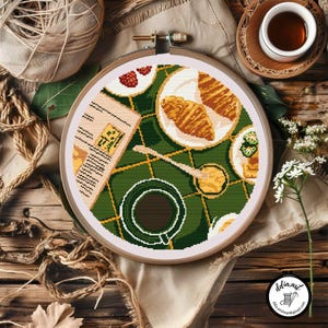 Può includere: Ricamo a punto croce in un telaio di legno raffigurante una scena di colazione. Una tovaglia verde a scacchi contiene una tazza di caffè, croissant e un giornale. La tavolozza dei colori comprende verde, marrone, bianco e giallo.
