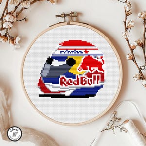Può includere: Un disegno a punto croce di un casco da corsa in un telaio da ricamo in legno. Il casco presenta il logo Red Bull in rosso, giallo e bianco su uno sfondo blu e grigio. Il disegno è su tessuto bianco.
