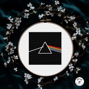 Può includere: Un ricamo a punto croce in un cerchio bianco presenta un quadrato nero con un triangolo bianco e uno spettro arcobaleno. Il disegno è circondato da fiori bianchi e posto su uno sfondo verde scuro. La firma dell'artista è in basso a destra.