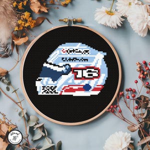 May include: Cross-stitch embroidery of a racing helmet in a wooden hoop. The helmet design features white, blue, and red colors, with the number 16 prominently displayed. The background is black, and the hoop is surrounded by floral elements.