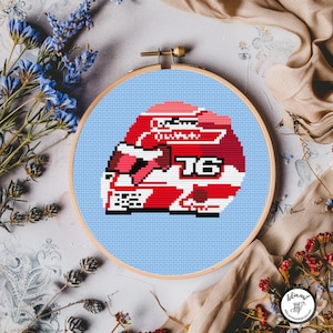 May include: Cross-stitch design featuring a red and white racing helmet with the number 16, set against a light blue backdrop. The artwork is displayed in a wooden embroidery hoop, with a pixelated aesthetic.