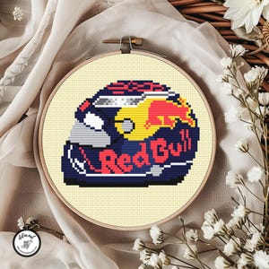 Può includere: Un ricamo a punto croce di un casco da corsa Red Bull in un telaio di legno. Il casco presenta il logo Red Bull in rosso e giallo su sfondo blu navy. Il testo "Red Bull" è ben visibile.