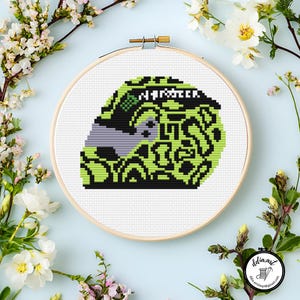 Op de afbeelding: Een kruissteekontwerp van een helm met het woord "Monster" in het wit. De helm heeft een zwart en neon groen patroon, met een grijs vizier. Het ontwerp is geplaatst in een houten borduurring.