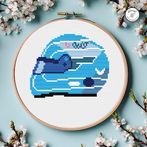 May include: A cross-stitch embroidery hoop with a blue helmet design. The helmet, in shades of blue, has a visor. The embroidery is set against a white fabric background within a wooden hoop. The design is pixelated.