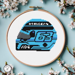May include: Cross-stitch embroidery of a racing helmet in a wooden hoop. The helmet is light blue with black accents and the number 63. The text "+S+U+E+R+S" is above the helmet visor. The background is light blue with white flowers.