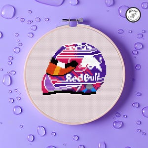 May include: A cross-stitch embroidery of a Red Bull racing helmet in a wooden hoop. The helmet design features the Red Bull logo in white and the text "Red Bull" in red. The color palette includes shades of purple, pink, orange, and white.