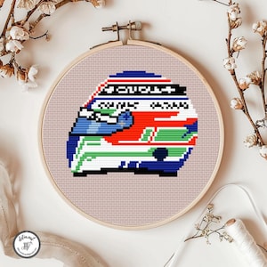 May include: A cross-stitch embroidery project featuring a pixelated racing helmet design in red, white, blue, and green colours. The helmet is centred within a circular wooden embroidery hoop, set against a light pink fabric background.