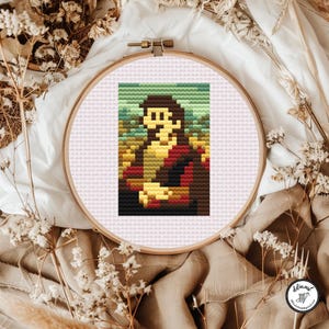 Può includere: Un telaio da ricamo circolare in legno contenente un motivo a punto croce di una figura pixelata con capelli castani, che indossa un indumento rosso scuro, su uno sfondo paesaggistico verde e giallo su un tessuto a griglia rosa chiaro.