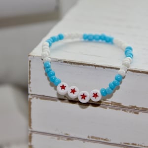 Peut inclure: Un bracelet en perles bleu et blanc avec trois étoiles rouges. Le bracelet est sur une surface en bois blanc.