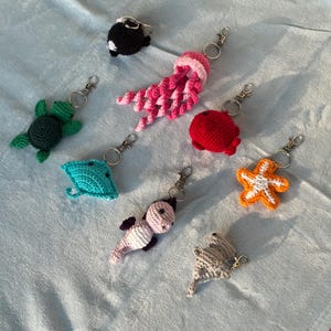 May include: A collection of eight crocheted sea creature keychains including a black whale, a pink jellyfish, a green turtle, a red fish, a turquoise stingray, an orange starfish, a light pink seahorse, and a grey stingray, each with a metal ring.