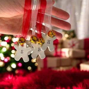 Peut inclure: Ornements blancs en forme de bonhomme en pain d'épices avec des cloches dorées et des rubans rouges et blancs. Chaque ornement porte une lettre : S, B et H. Les ornements sont tenus devant un sapin de Noël et des cadeaux.