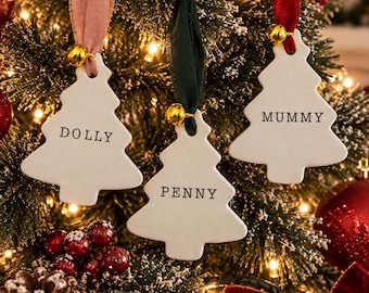 Könnte beinhalten: Drei weiße Weihnachtsbaumanhänger aus Keramik in Tannenbaumform mit glatten Oberflächen, jeweils mit einer kleinen goldenen Glocke und einem farbigen Band. Die Anhänger tragen die schwarze Aufschrift DOLLY, PENNY und MUMMY an einem Tannenzweig.