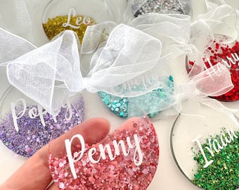 May include: Clear, round ornaments with glitter and names in white script. The ornaments are decorated with various colors of glitter, including gold, purple, teal, red, and green. Each ornament is tied with a white ribbon.