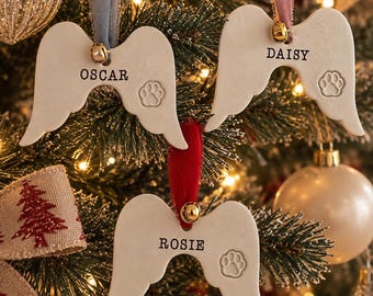 May include: Three white ceramic angel wing ornaments, each featuring a stamped paw print and a name: OSCAR, DAISY, and ROSIE. Each ornament hangs from a coloured velvet ribbon with a small gold bell attached at the top centre of the wing shape.