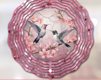 May include: A pink metal wind spinner with a design of two hummingbirds perched on a branch with pink blossoms. The wind spinner has a circular shape with multiple layers of metal.