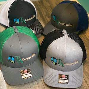 Könnte beinhalten: Vier graue und schwarze Trucker-Caps mit einem grün und blau gestickten Logo, auf dem "TA Construction" steht. Das Logo zeigt eine stilisierte Ölbohrplattform. Die Caps haben ein schwarzes Netzrücken und ein Richardson-Label.