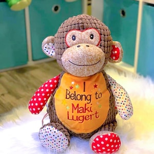 May include: A brown plush monkey with red and white polka dot accents. The monkey is wearing a yellow bib with the text "I Belong to Maki Lugert" embroidered on it.