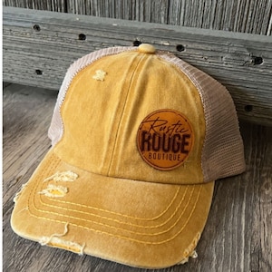 Puede incluir: Una gorra de camionero desgastada de color amarillo mostaza con un parche de cuero marrón que dice "Rustic Rouge Boutique".
