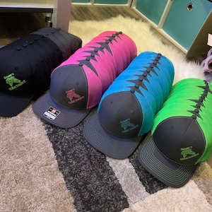 May include: A stack of black, pink, blue, and green mesh trucker hats with black bills. Each hat has a black embroidered logo that says "Shane's Guide Service" with a fish graphic.