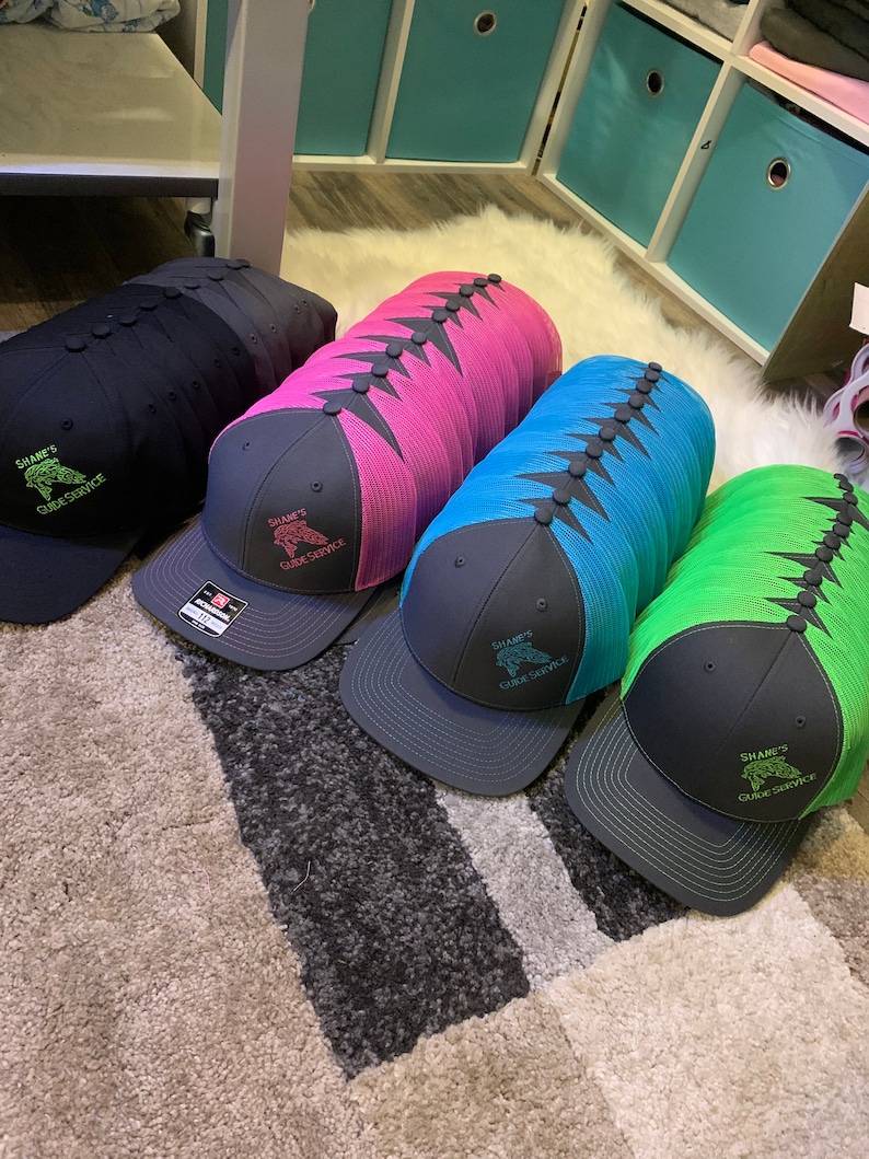 May include: A stack of black, pink, blue, and green mesh trucker hats with black bills. Each hat has a black embroidered logo that says "Shane's Guide Service" with a fish graphic.