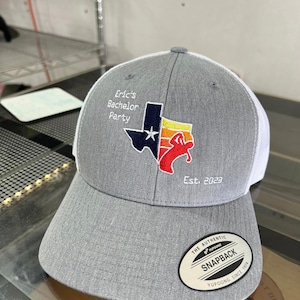 May include: Gray and white baseball cap with a Texas-shaped patch featuring a red, white, and blue design. The patch includes a star and the text "Eric's Bachelor Party Est. 2023". The cap has a white mesh back and a black "The Authentic Yupoong Snapback Yupoong Since 1974" label.