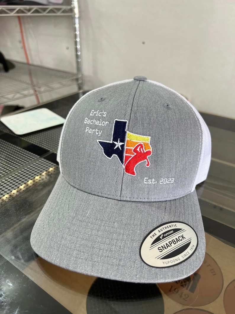 May include: Gray and white baseball cap with a Texas-shaped patch featuring a red, white, and blue design. The patch includes a star and the text "Eric's Bachelor Party Est. 2023". The cap has a white mesh back and a black "The Authentic Yupoong Snapback Yupoong Since 1974" label.