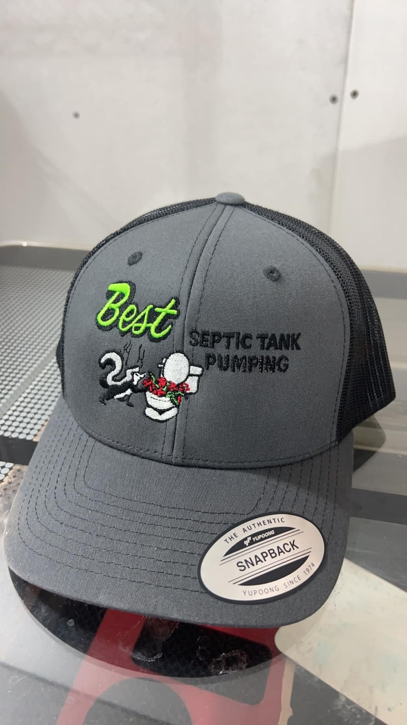 May include: A gray and black trucker hat with a white embroidered design of a cartoon skunk holding a toilet plunger. The text "Best Septic Tank Pumping" is embroidered in black on the hat. The hat has a black mesh back and a white "Snapback" label.