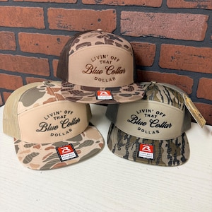 Könnte beinhalten: Drei Flat-Bill-Snapback-Hüte mit dem Text "LIVIN' OFF THAT Blue Collar DOLLAR". Die Hüte haben Tarnmuster in Braun-, Beige- und Grüntönen, Mesh-Rücken und eine beige Frontplatte.
