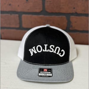 Könnte beinhalten: Schwarze, weiße und graue Trucker-Cap mit dem Wort "CUSTOM" in Weiß gestickt. Die Cap hat einen grauen Schirm, eine schwarze Front und eine weiße Mesh-Rückseite. Die Richardson 112 Trucker Cap ist verstellbar.