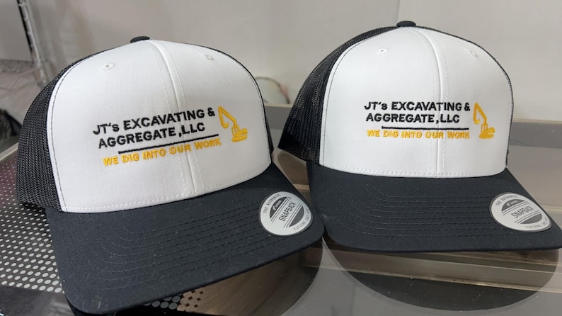 May include: Two white and black trucker hats with a yellow embroidered logo that says "JT's Excavating & Aggregate, LLC" and "We Dig Into Our Work".