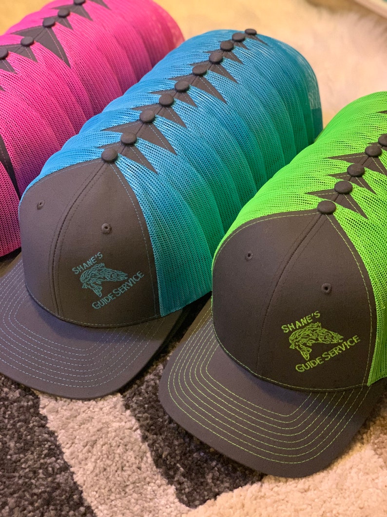 May include: Three black and gray trucker hats with mesh back panels. The hats have different colored mesh panels: pink, blue, and green. Each hat has a black embroidered logo that says "Shane's Guide Service" with a fish graphic.