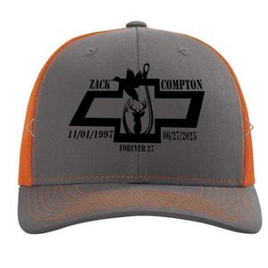 May include: A gray and orange trucker hat with a black graphic design. The design includes the name "ZACK COMPTON", dates, and a deer silhouette. The hat has a curved brim and a mesh back.