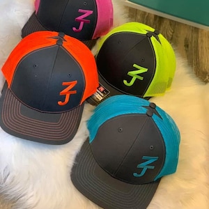 May include: Four black trucker hats with different colored mesh panels. The hats have a white embroidered "ZJ" logo on the front. The mesh panels are pink, green, orange, and blue.