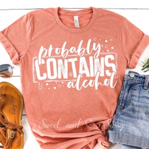 Puede incluir: Una camiseta de color coral con el texto "probably contains alcohol" en letras blancas con un aspecto desgastado. La camiseta está hecha de un material suave y cómodo.