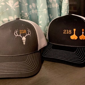 May include: Two black and white trucker hats with mesh panels. One hat has a white deer skull with antlers and the number 218 embroidered on the front. The other hat has two orange paddles and the number 218 embroidered on the front.