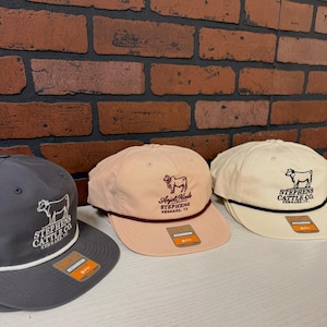 以下が含まれることがあります： 3つの野球帽が並んでおり、それぞれ異なる色とデザインです。帽子はグレー、ピーチ、クリーム色です。各帽子には牛のロゴと「Stephens Cattle Co. Kennard, TX」または「Anjoli Nicole Stephens Kennard, TX」の文字が入っています。
