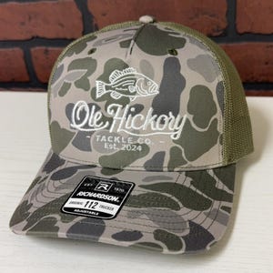 以下が含まれることがあります： 白地に「Ole Hickory Tackle Co. Est. 2024」と刺繍されたロゴと、白く刺繍された魚が描かれた、迷彩柄の緑と茶色のトラッカーハット。リチャードソン112製です。