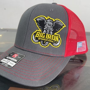 May include: Gray and red trucker hat with a black and yellow embroidered logo that says "Big Iron International". The logo features an elephant with tusks. The hat has a small American flag on the side.