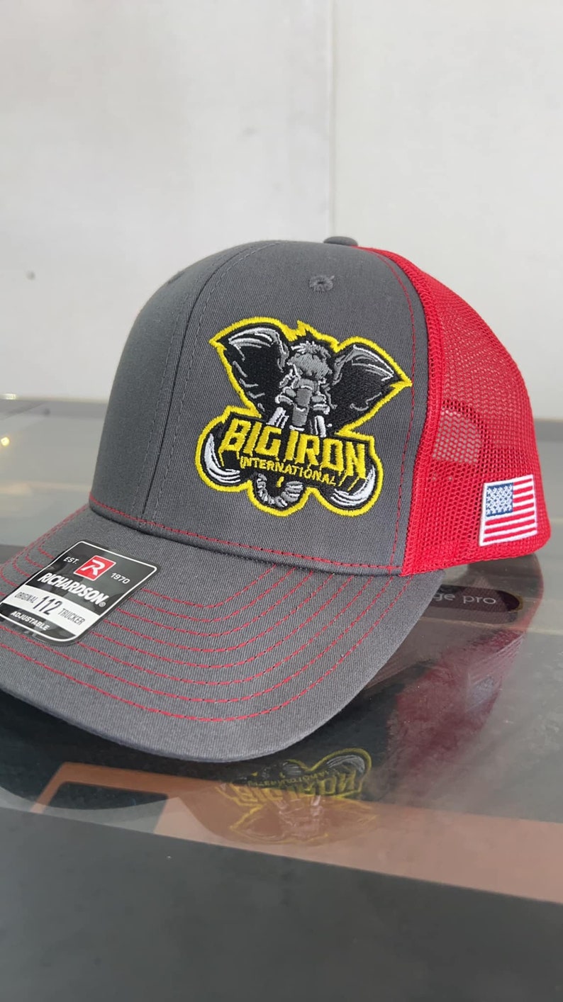 May include: Gray and red trucker hat with a black and yellow embroidered logo that says "Big Iron International". The logo features an elephant with tusks. The hat has a small American flag on the side.