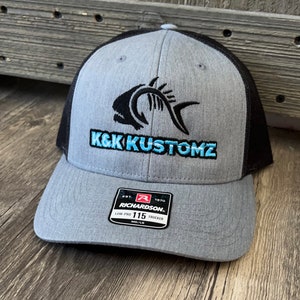 May include: Grey and black trucker cap featuring a fish skeleton design and the teal text "K&K Kustomz". The cap has a curved peak and a mesh back. The Richardson 115 Low Pro Trucker cap is a MD/LG size.