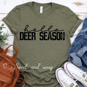 May include: Olive green t-shirt with the words "hello DEER SEASON" in black script and block letters. The shirt is styled with a brown backpack, white sneakers, and a denim jacket. The text "Sweet and sassy" is also visible.