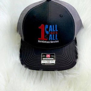 May include: Black and gray trucker hat with a red and blue embroidered logo that says "1 Call Thats All Handyman Service". The hat is adjustable and has a Richardson label.