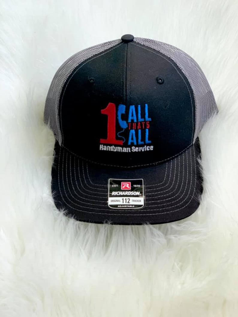 May include: Black and gray trucker hat with a red and blue embroidered logo that says "1 Call Thats All Handyman Service". The hat is adjustable and has a Richardson label.