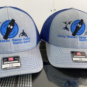 May include: Two gray and blue trucker hats with a black and blue logo that says "Dirty Water Game Calls Quality Quack" and "DW" in a circle. The hats are both Richardson brand and have a size tag that says "112".
