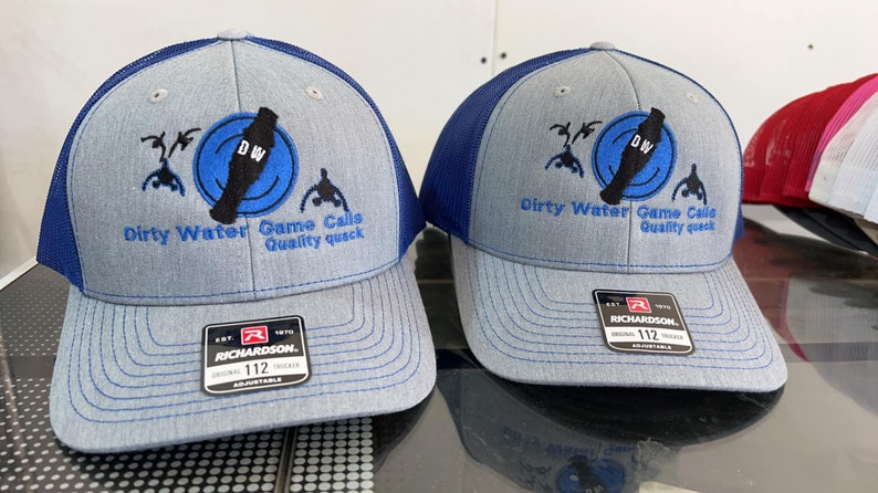 May include: Two gray and blue trucker hats with a black and blue logo that says "Dirty Water Game Calls Quality Quack" and "DW" in a circle. The hats are both Richardson brand and have a size tag that says "112".